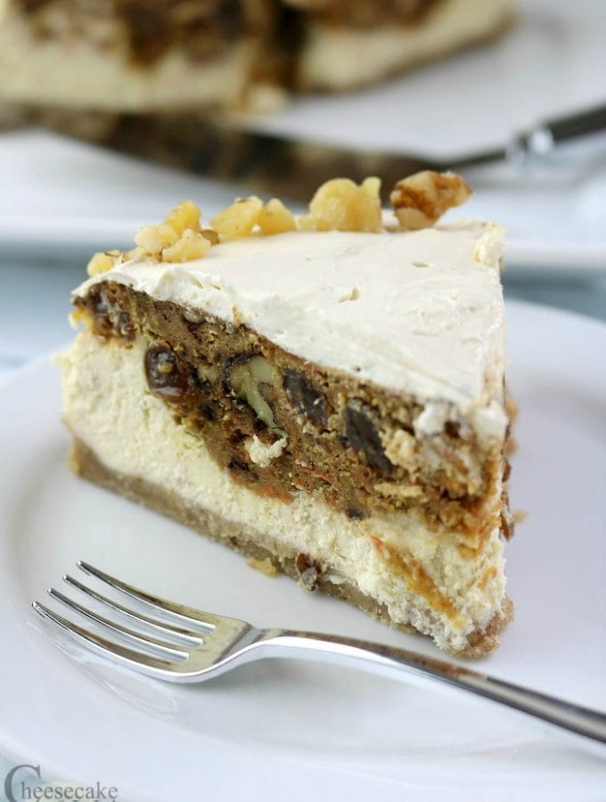 Slice of carrot cake cheesecake on a white plate with a fork. Rest of cheesecake in background.
