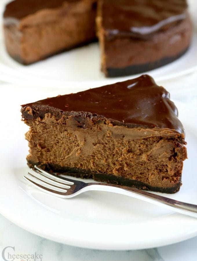 Slice of Triple Chocolate Cheesecake on a white plate with a fork. Rest of cheesecake in the background.