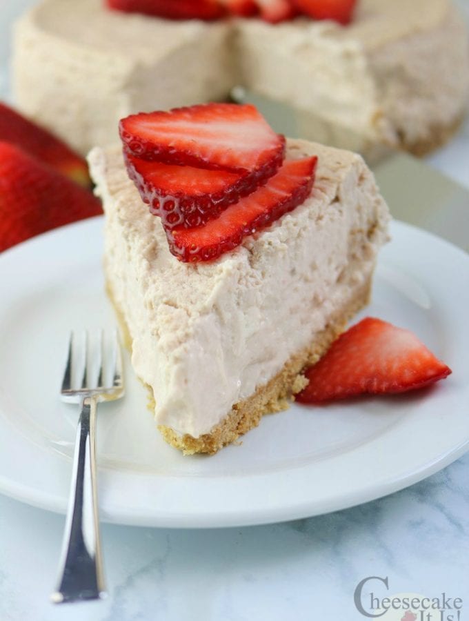 Slice of Strawberries And Cream Cheesecake on a white plate with a fork the rest of the cheesecake in background.