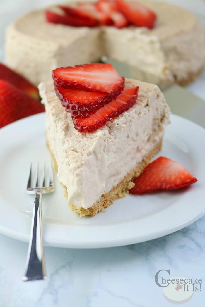 Slice of Strawberries And Cream Cheesecake on a white plate with a fork the rest of the cheesecake in background.