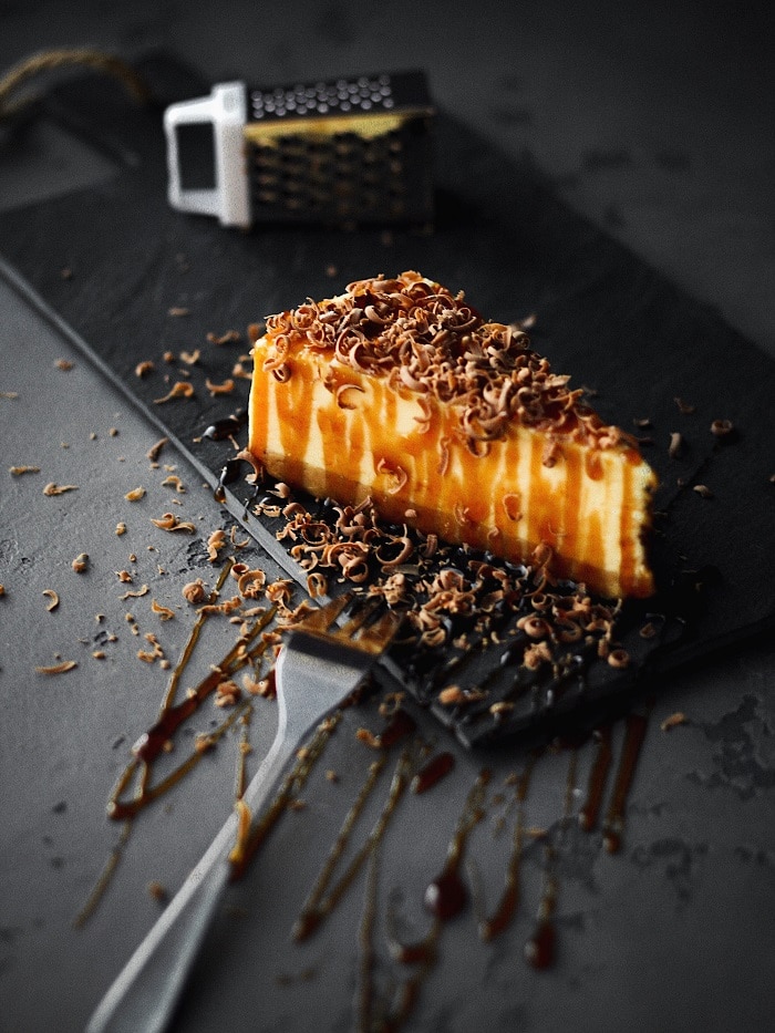 Slice of cheesecake on slate covered in sauce and chocolate shavings.