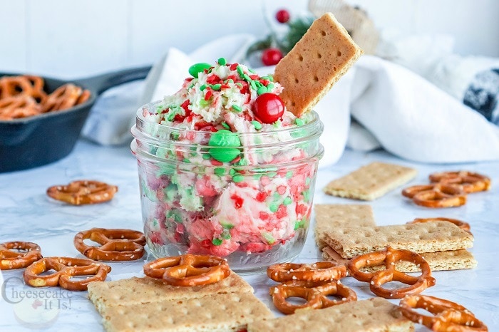 Glass jar full of sugar cookie cheesecake dip with red and green M&M's in it. Pretzels and graham crackers all around the jar