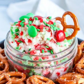 Glass jar full of sugar cookie cheesecake dip with red and green M&M's in it. Pretzels all around the jar