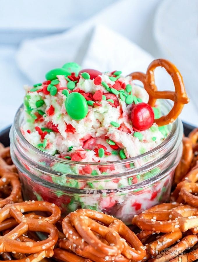 Glass jar full of sugar cookie cheesecake dip with red and green M&M's in it. Pretzels all around the jar