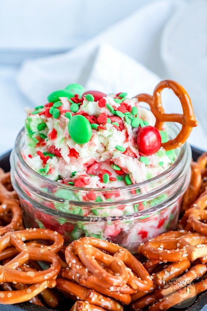 Glass jar full of sugar cookie cheesecake dip with red and green M&M's in it. Pretzels all around the jar