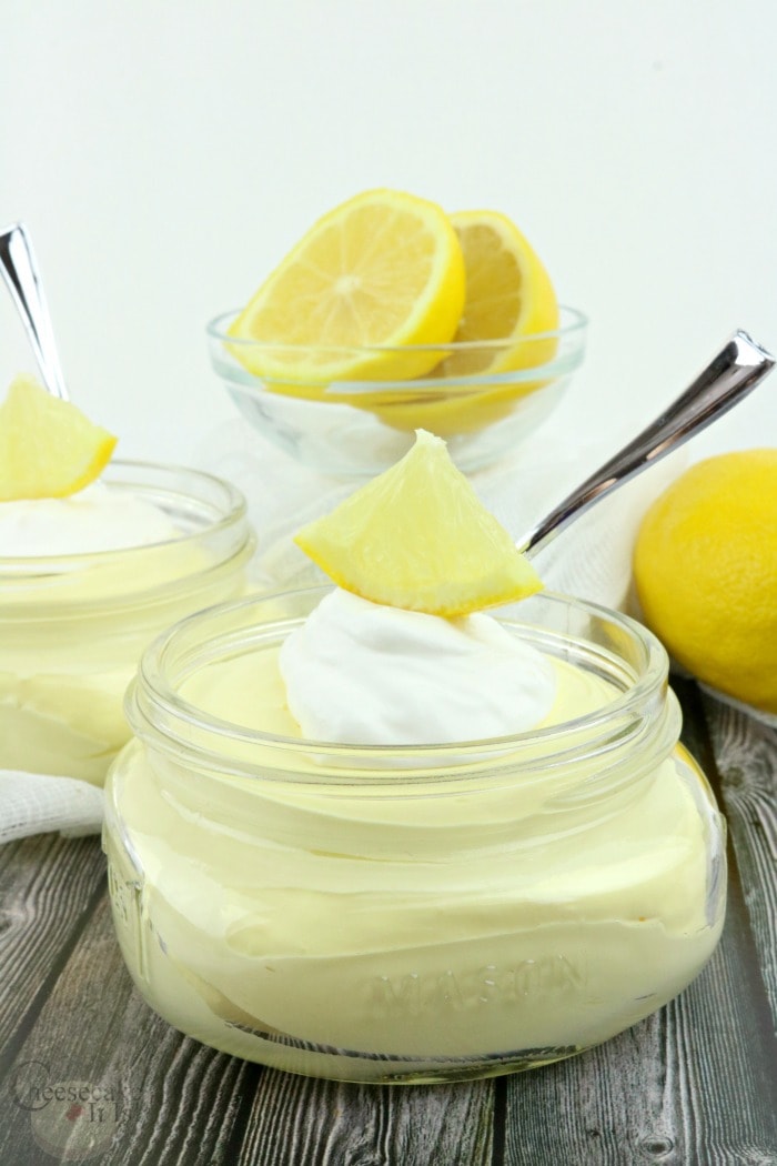 Jar of lemon cheesecake fluff topped with whipped cream and a piece of lemon. Dish of sliced lemon in background