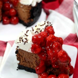 Slice of black forest cheesecake on white plate with rest of cheesecake in background