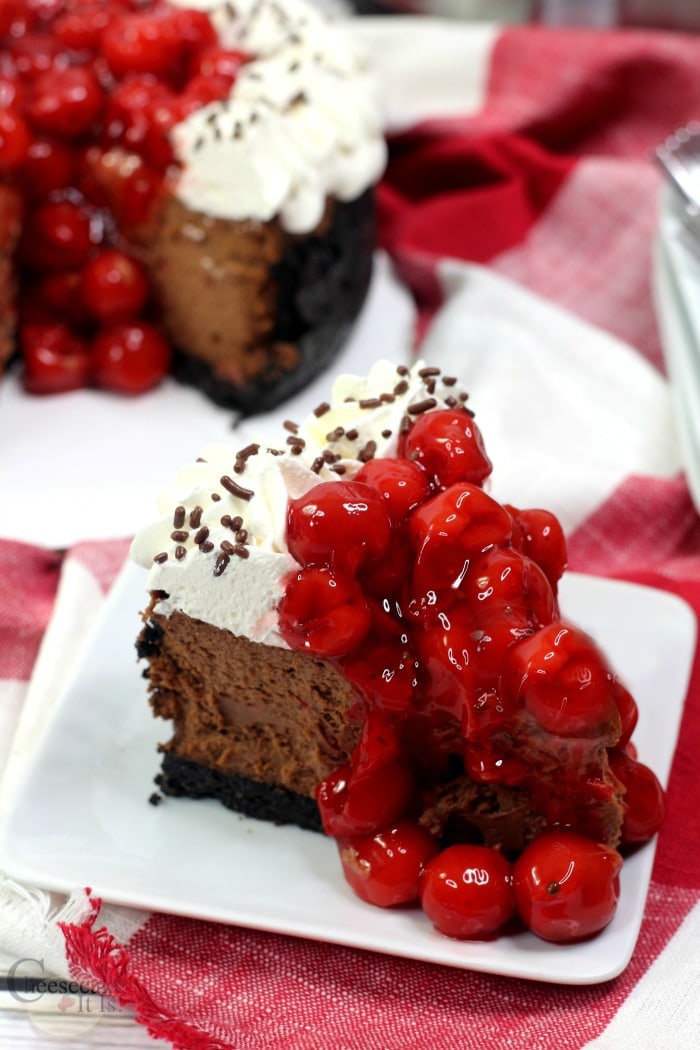 Slice of black forest cheesecake on white plate with rest of cheesecake in background