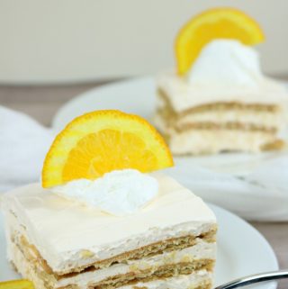 Two slices of creamsicle cheesecake icebox cake both on white plates with a half orange slice on top