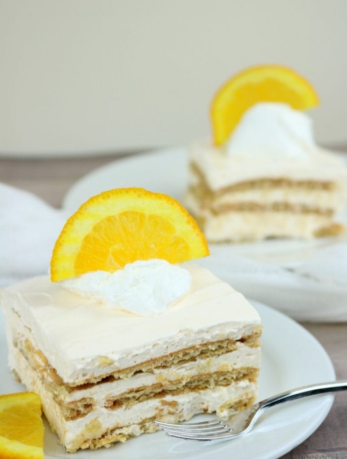 Two slices of creamsicle cheesecake icebox cake both on white plates with a half orange slice on top