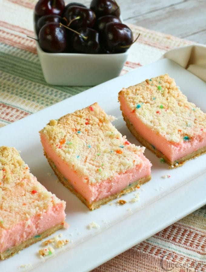 3 cheery cheesecake bars on white plate with whole cherries in background