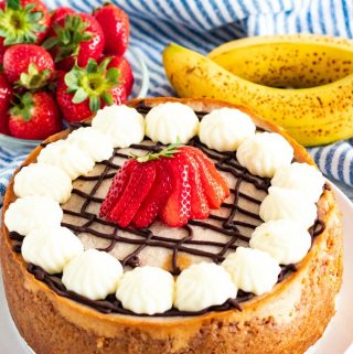 Whole banana split cheesecake on white plate with strawberries and bananas in background