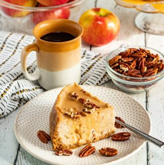 Slice of instant pot caramel apple cheesecake on white plate with coffee nuts and apples in background