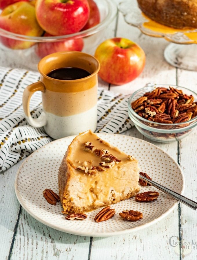 Slice of instant pot caramel apple cheesecake on white plate with coffee nuts and apples in background