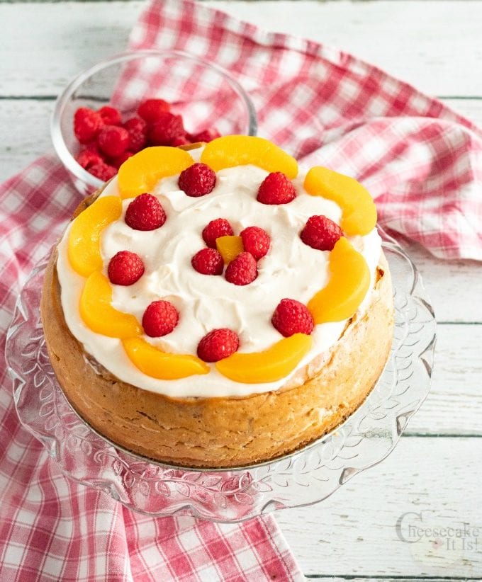 Whole Raspberry Peach Cheesecake on cake stand topped with fresh fruit and cream