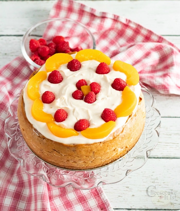 Whole Raspberry Peach Cheesecake on cake stand topped with fresh fruit and cream