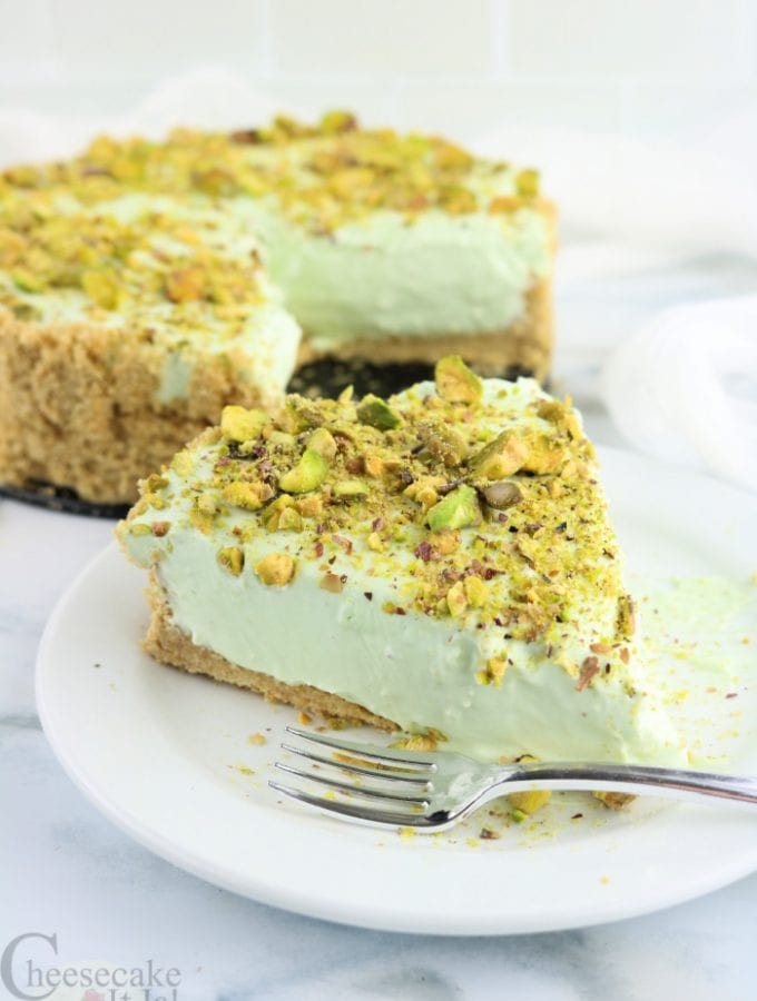 Slice of pistachio cheesecake on white plate with fork. Rest of whole cheesecake in background
