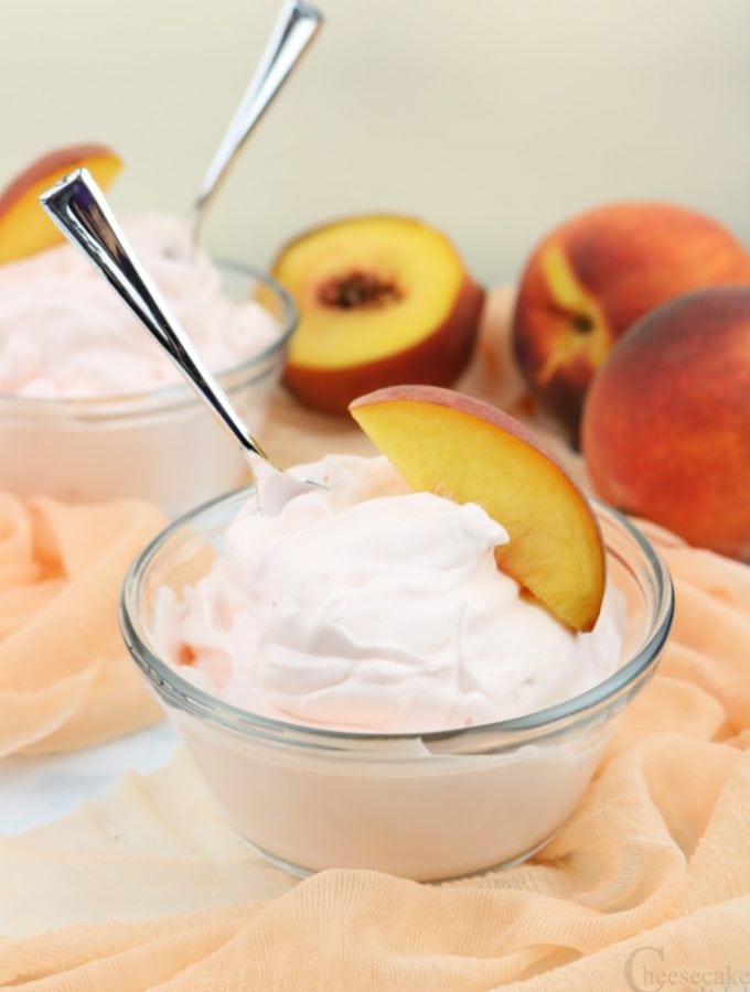 Small glass dish of peach cheesecake fluff with fresh peach slice on top and fresh peaches in background