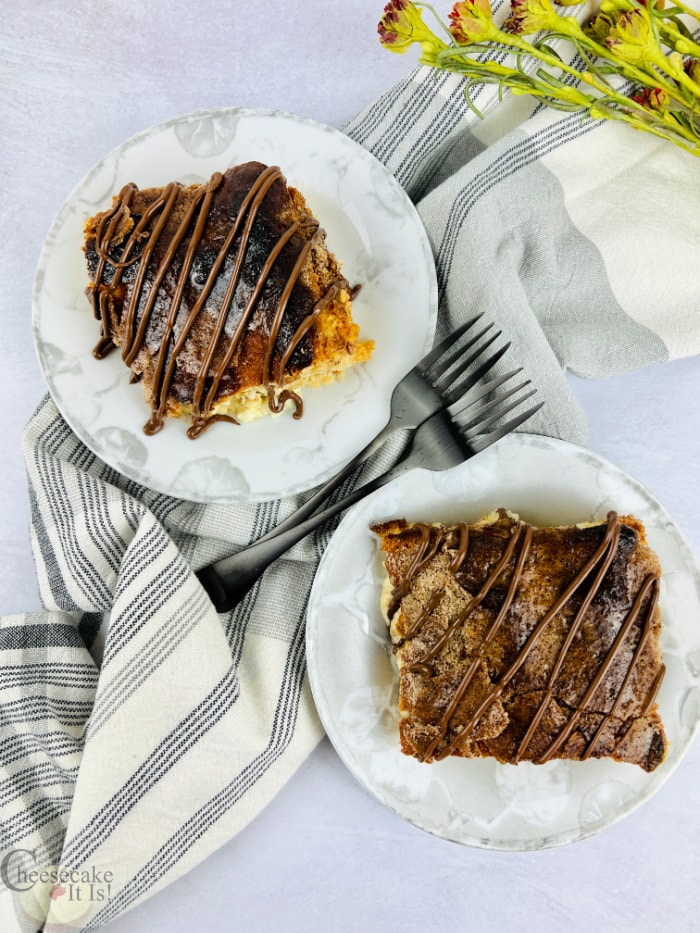Two slices of churro cheesecake on two white plates with dish towel and forks to side