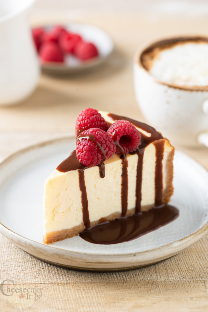 Slice of cheesecake on white plate sitting on wood. Cake topped with chocolate sauce and raspberries