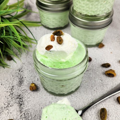 Small jar full of keto pistachio cheesecake fluff topped with whip cream and pistachio nuts. More full jars with lids stacked in background