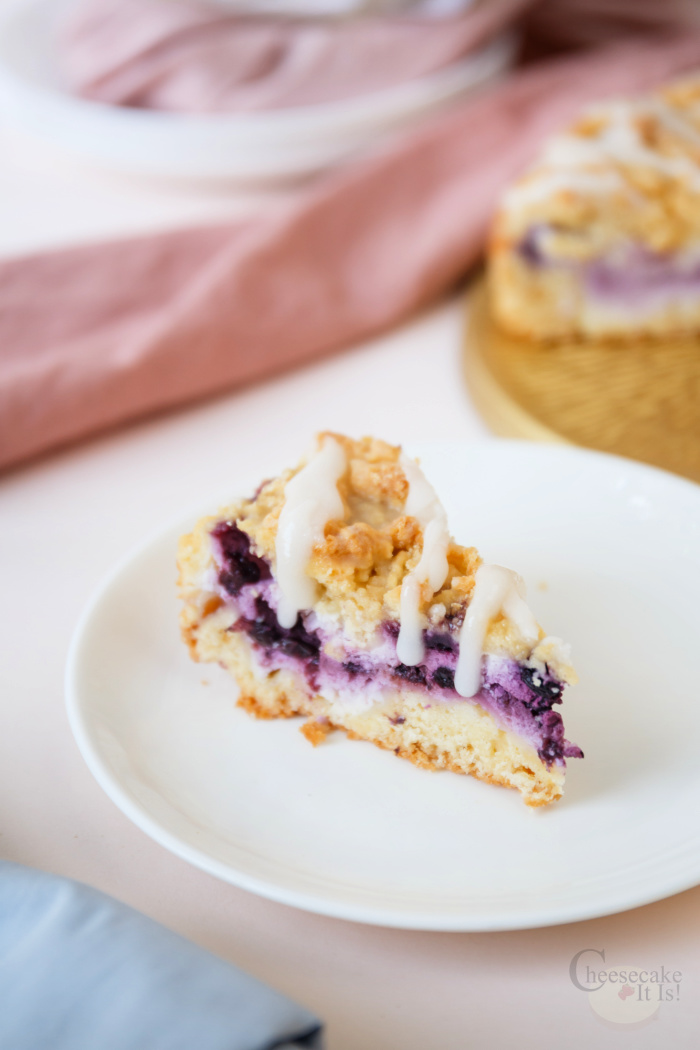 Slice of blueberry crumble cheesecake on white plate. Rest of cheesecake in background.