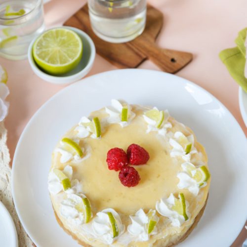 Whole key lime cheesecake on white plate topped with whip cream raspberries and lime slices