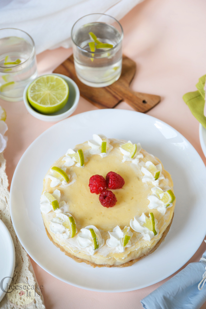 Whole key lime cheesecake on white plate topped with whip cream raspberries and lime slices