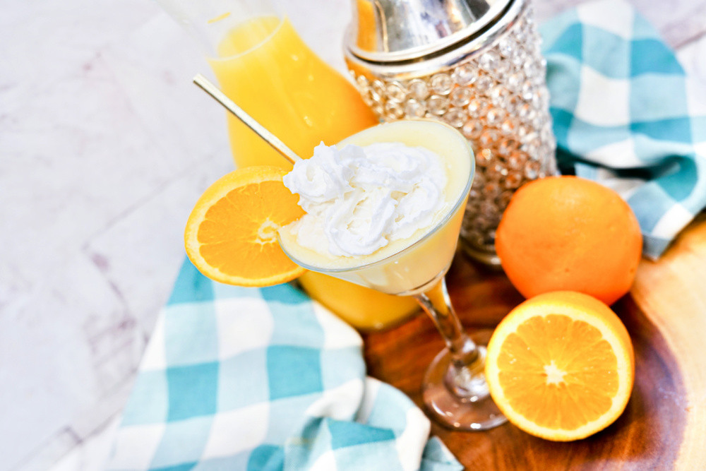 An tilted shot of a Cheesecake Orange Julius Martini in a glass topped with whipped cream and orange slice.