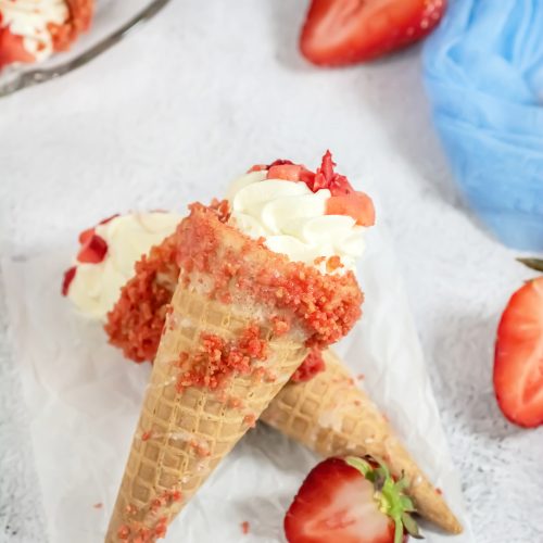 Two strawberry crunch cheesecake cones criss crossed on piece of parchment paper with cut strawberries to the side