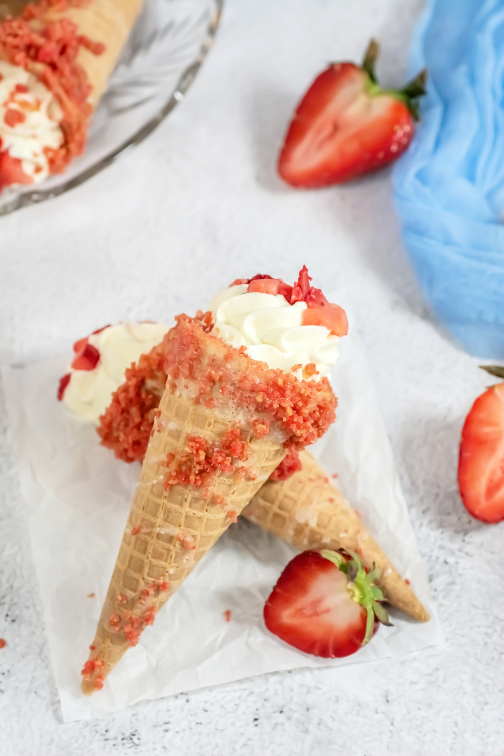 Two strawberry crunch cheesecake cones criss crossed on piece of parchment paper with cut strawberries to the side